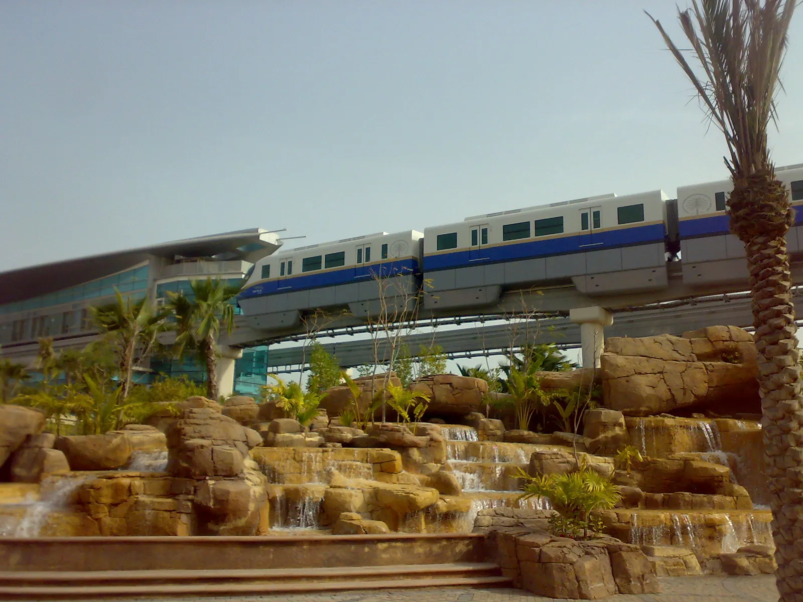 A blue-and-white Palm Jumeirah monorail train running along its elevated track past palms and a rock-and-waterfall garden, with Gateway station behind it