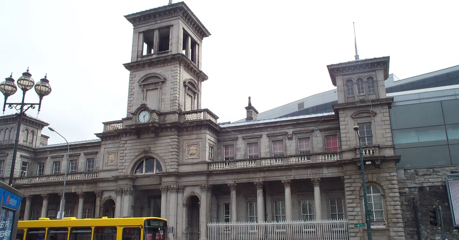 The granite front of Dublin Connolly Station on Amiens Street, a long Italianate range of arched windows with a square clock tower rising at its centre and a yellow city bus passing below