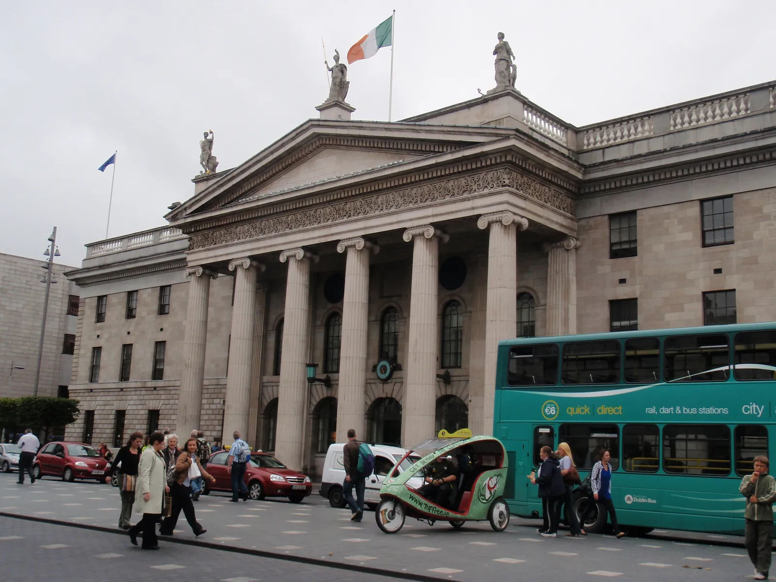 The General Post Office on O'Connell Street in Dublin, a grey stone portico of six tall Ionic columns under a pediment with statues on top and the Irish tricolour flying above, with buses and pedestrians in front