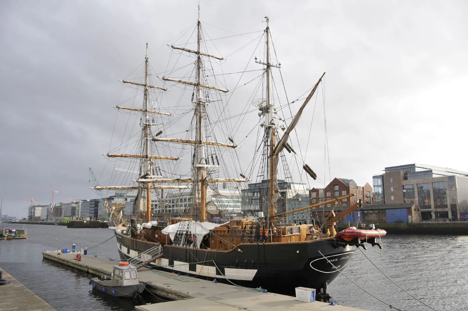 The Jeanie Johnston tied up on the Liffey in Dublin, a black-hulled three-masted sailing ship with its rigging up and the glass and brick blocks of the docklands behind it
