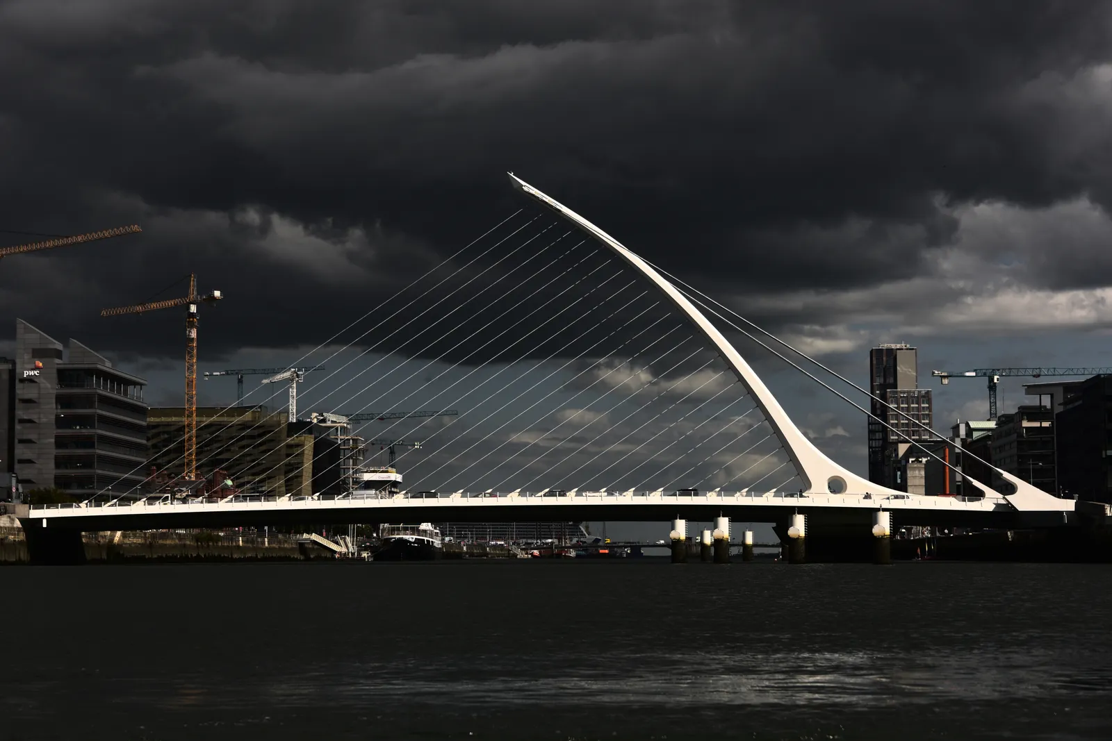 The Samuel Beckett Bridge across the Liffey in Dublin, a white curved spar leaning back over the water with steel cables fanning down to the deck like the strings of a harp, under a dark sky