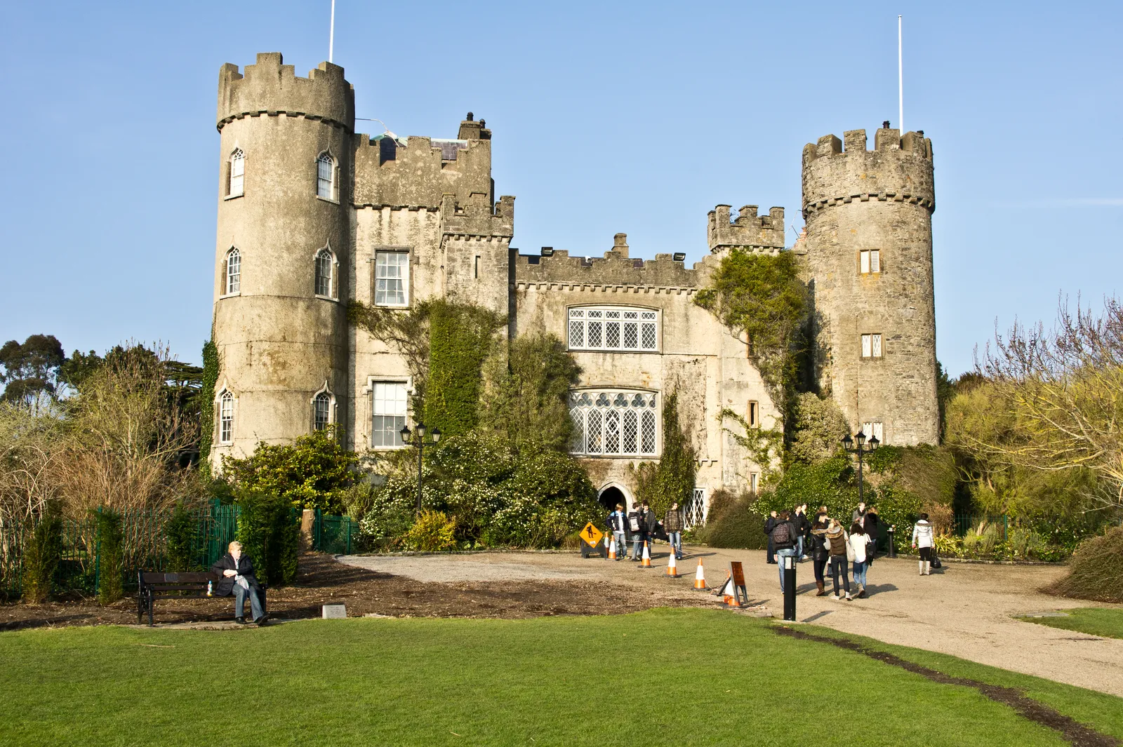 Malahide Castle in County Dublin seen across its lawns, a pale stone house with two round battlemented towers at its corners and ivy climbing the front, with visitors walking on the gravel below