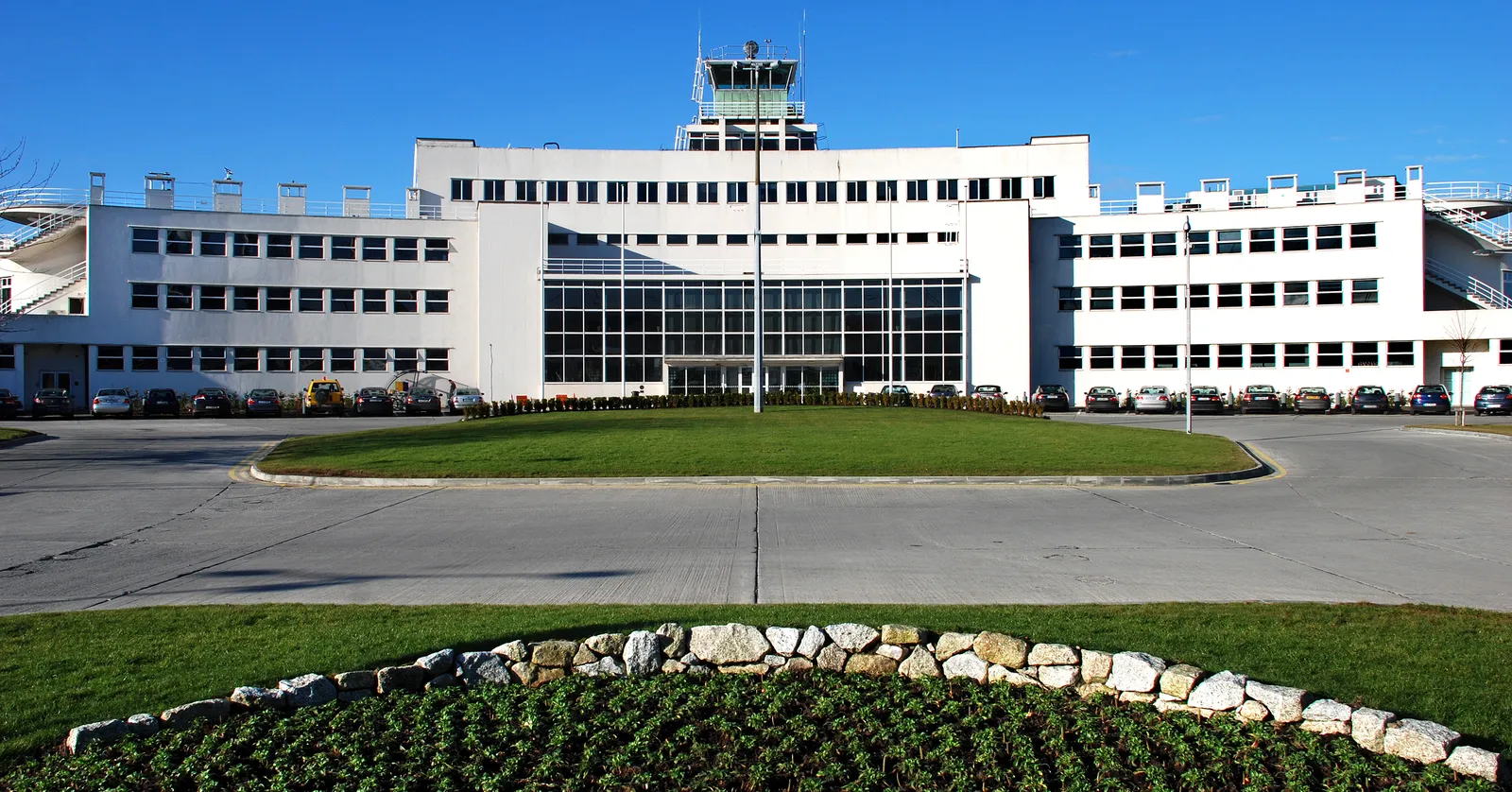 The original 1940 terminal building at Dublin Airport, a long white curved modernist block of horizontal window bands with a glazed control tower on the roof, a lawn and flowerbed in front of it