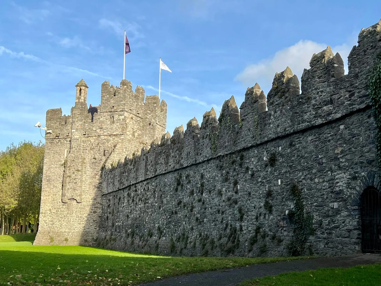The curtain wall of Swords Castle in County Dublin, a long battlemented grey stone wall running along a mown lawn to a square tower with flags flying from it, under a blue sky
