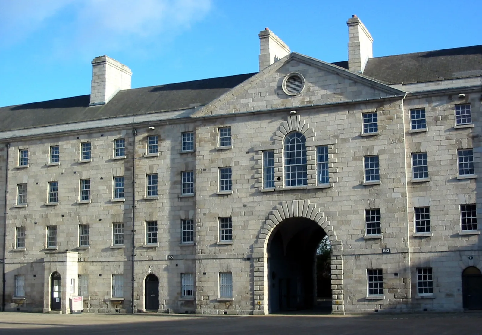 A range of Collins Barracks in Dublin under a blue sky, four storeys of plain grey granite with rows of small windows and a wide arched carriage entrance through the middle