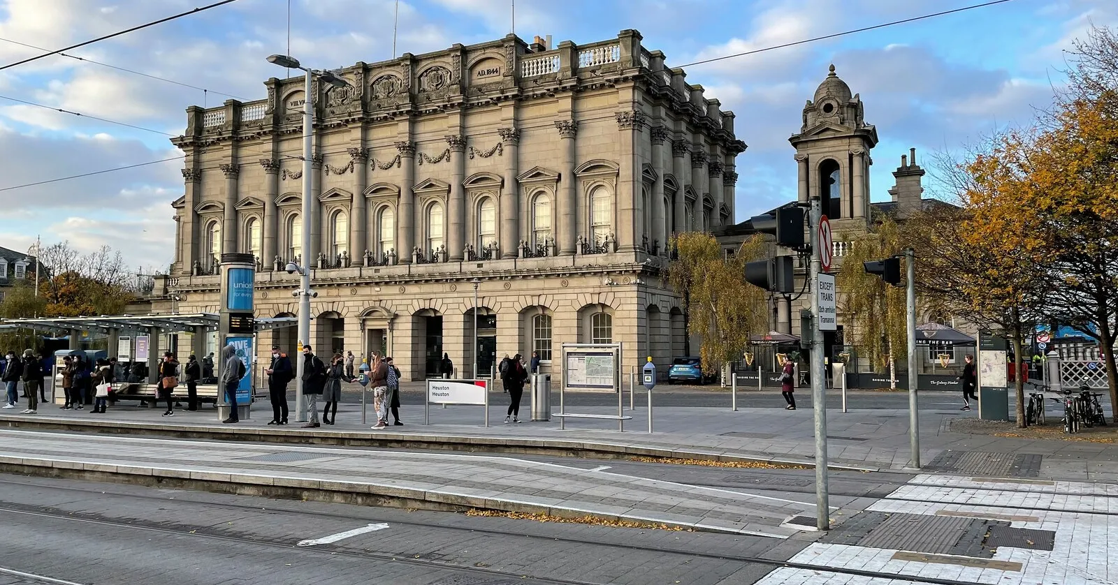 The front of Dublin Heuston Station in autumn sunlight, a long carved limestone block in the style of an Italian palazzo with a bell tower at one end and passengers waiting at the tram stop in front