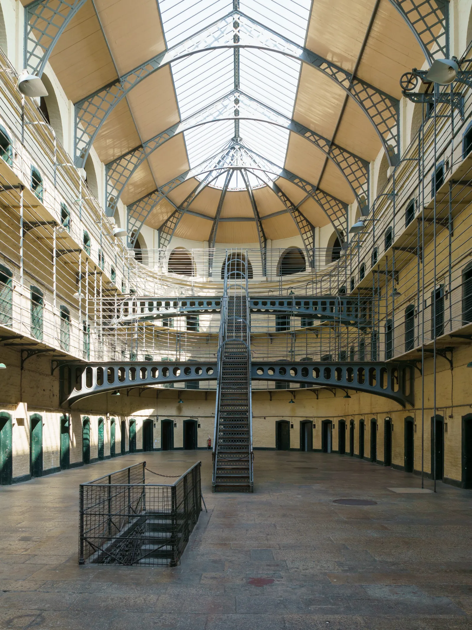 The east wing of Kilmainham Gaol, a tall barrel-vaulted hall of pale stone with three tiers of iron balconies and cell doors around it and a metal staircase climbing the middle of the floor