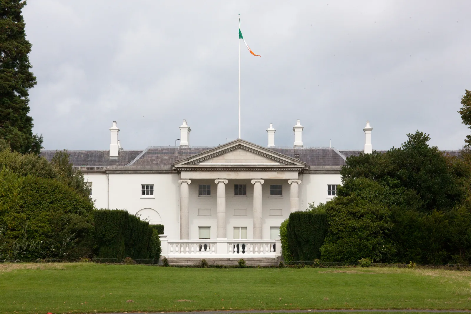 Áras an Uachtaráin in Dublin's Phoenix Park, a long white house with a columned portico and a balustraded terrace, the Irish tricolour flying from a mast above it and lawn in front