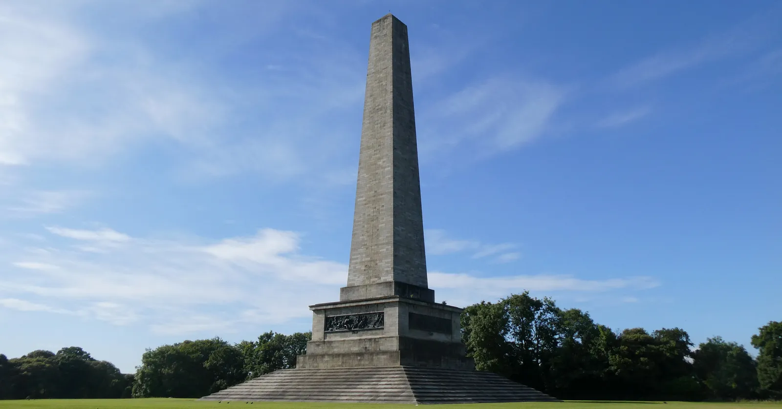 The Wellington Testimonial in Dublin's Phoenix Park, a tall grey granite obelisk on a stepped plinth standing alone on a wide expanse of mown grass under a blue sky