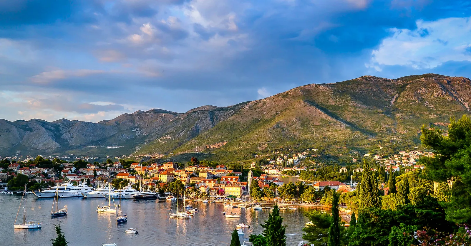 Cavtat's harbour in late afternoon light, terracotta roofs and a bell tower along the shore, yachts and small boats on flat water, and bare limestone mountains rising behind the town