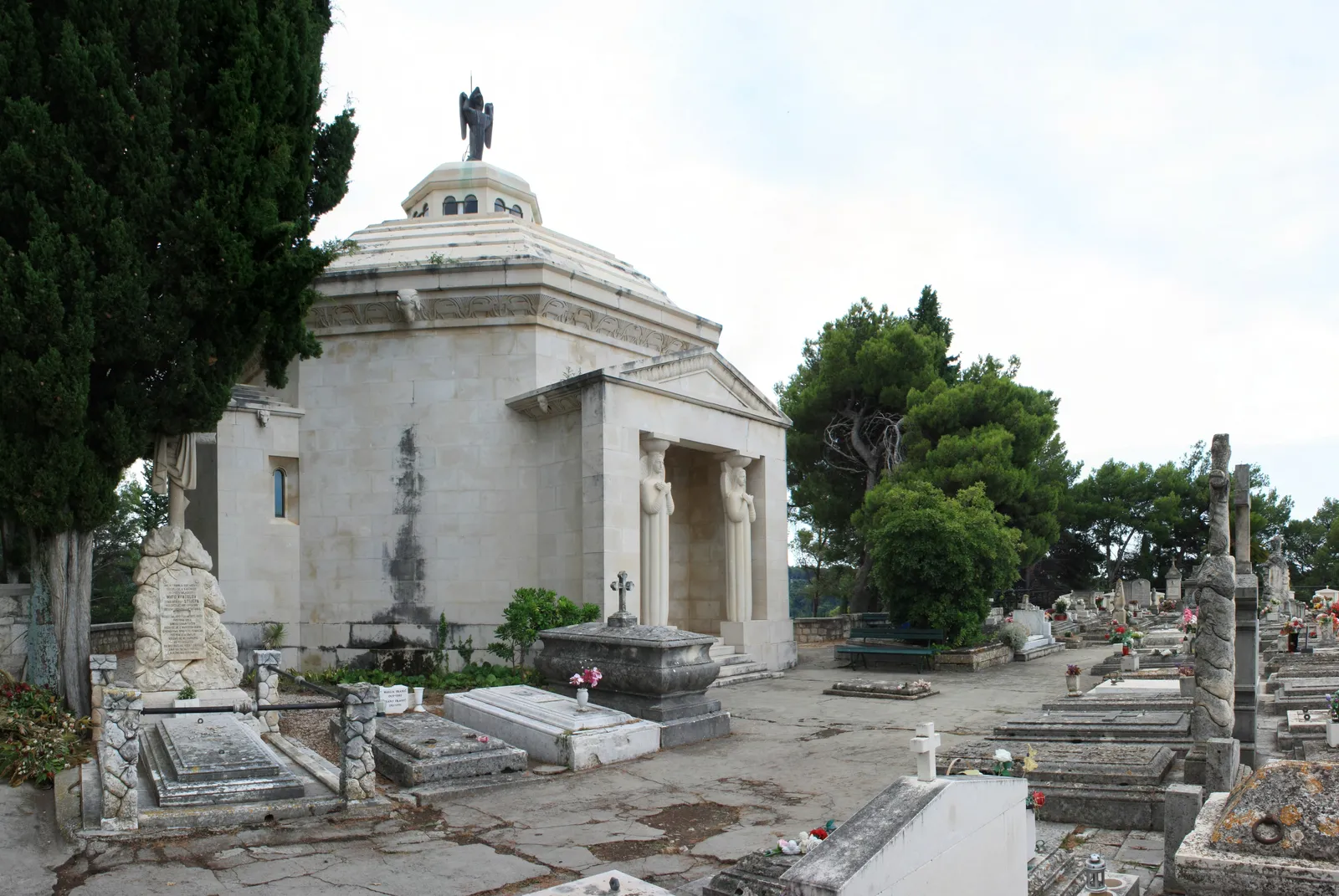 The Račić family mausoleum at St Roch cemetery above Cavtat, a domed white stone building with a columned porch and a figure on the roof, surrounded by cypresses and older graves