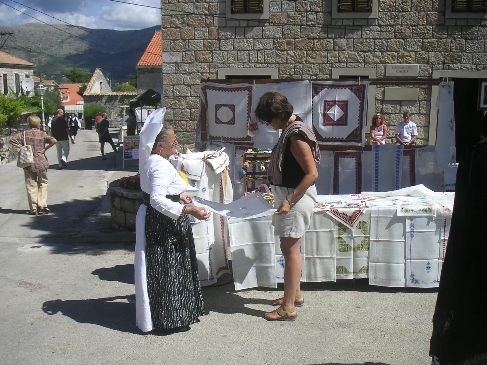 A woman in traditional Konavle dress showing embroidered white linen to a visitor at a village stall in Čilipi, with more stitched cloths hung on a display board behind and hills in the distance