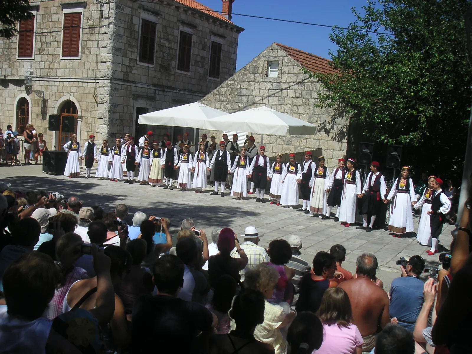 Dancers in white and black Konavle costume standing in a line across a stone village square in Čilipi, watched by a crowd of seated and standing spectators in front of old stone houses