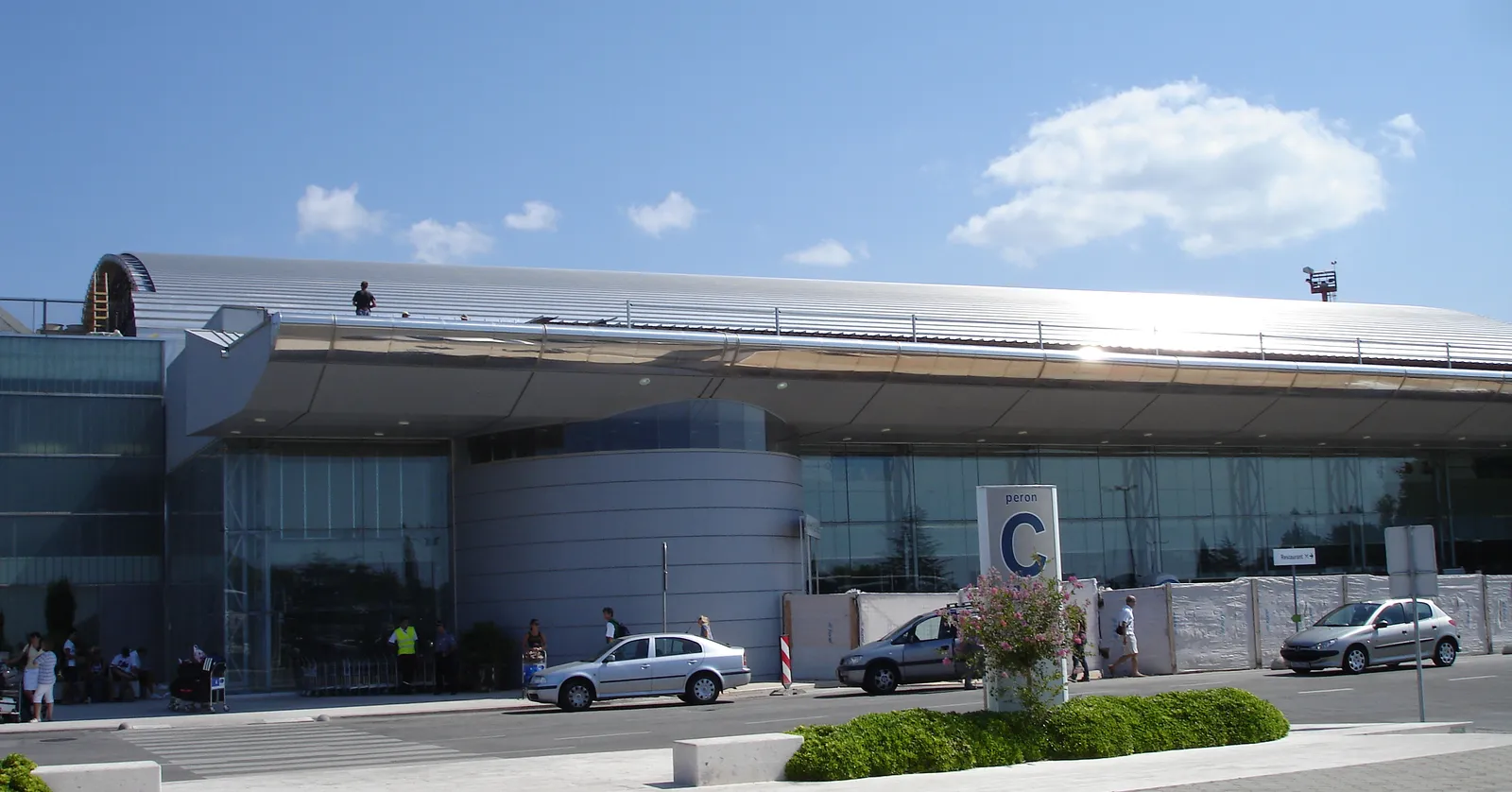 The terminal building at Dubrovnik airport under a bright sky, a long low block of glass and grey panelling beneath a sweeping metal roof, with cars pulled up on the forecourt outside