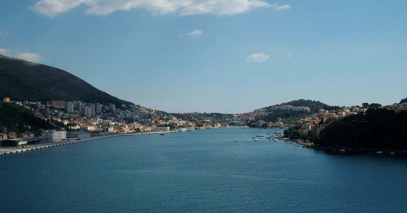 The long inlet of Gruž harbour in Dubrovnik seen from above, deep blue water running inland between two hillsides covered in pale apartment blocks, with the quays and moored boats lining the left-hand shore