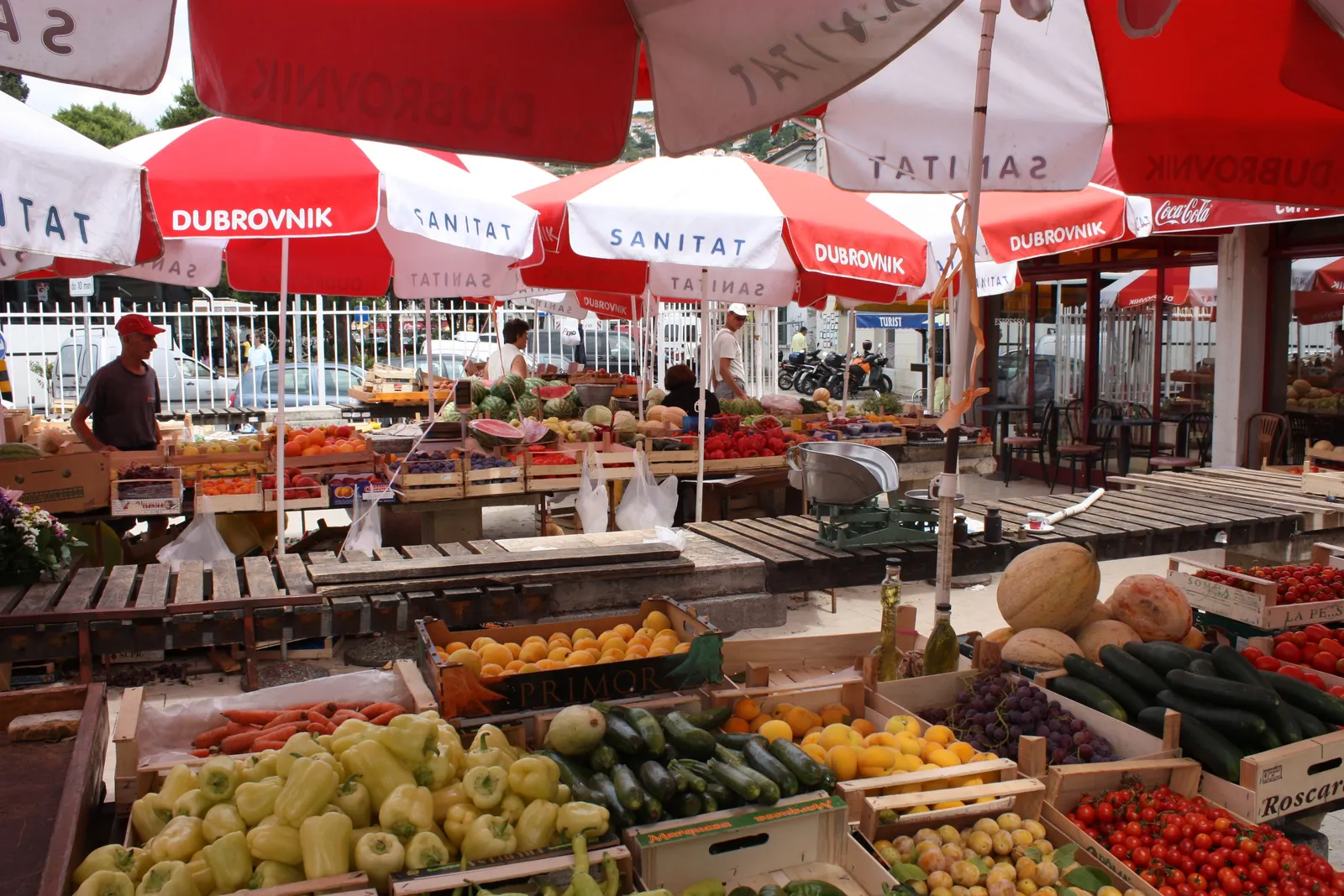 Open-air stalls at the Gruž market in Dubrovnik under red and white parasols, crates of peppers, tomatoes, cucumbers, melons and grapes stacked along the front of the tables