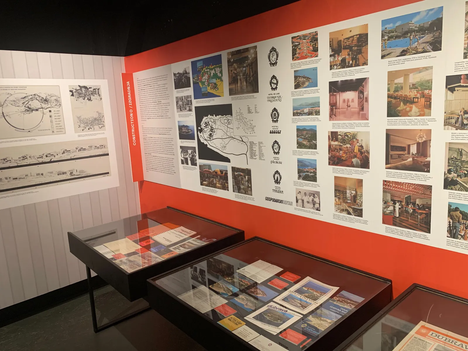 A gallery inside the Red History Museum in Dubrovnik, an orange wall covered in Yugoslav-era tourist posters and brochures with glass display cases of printed material in front of it