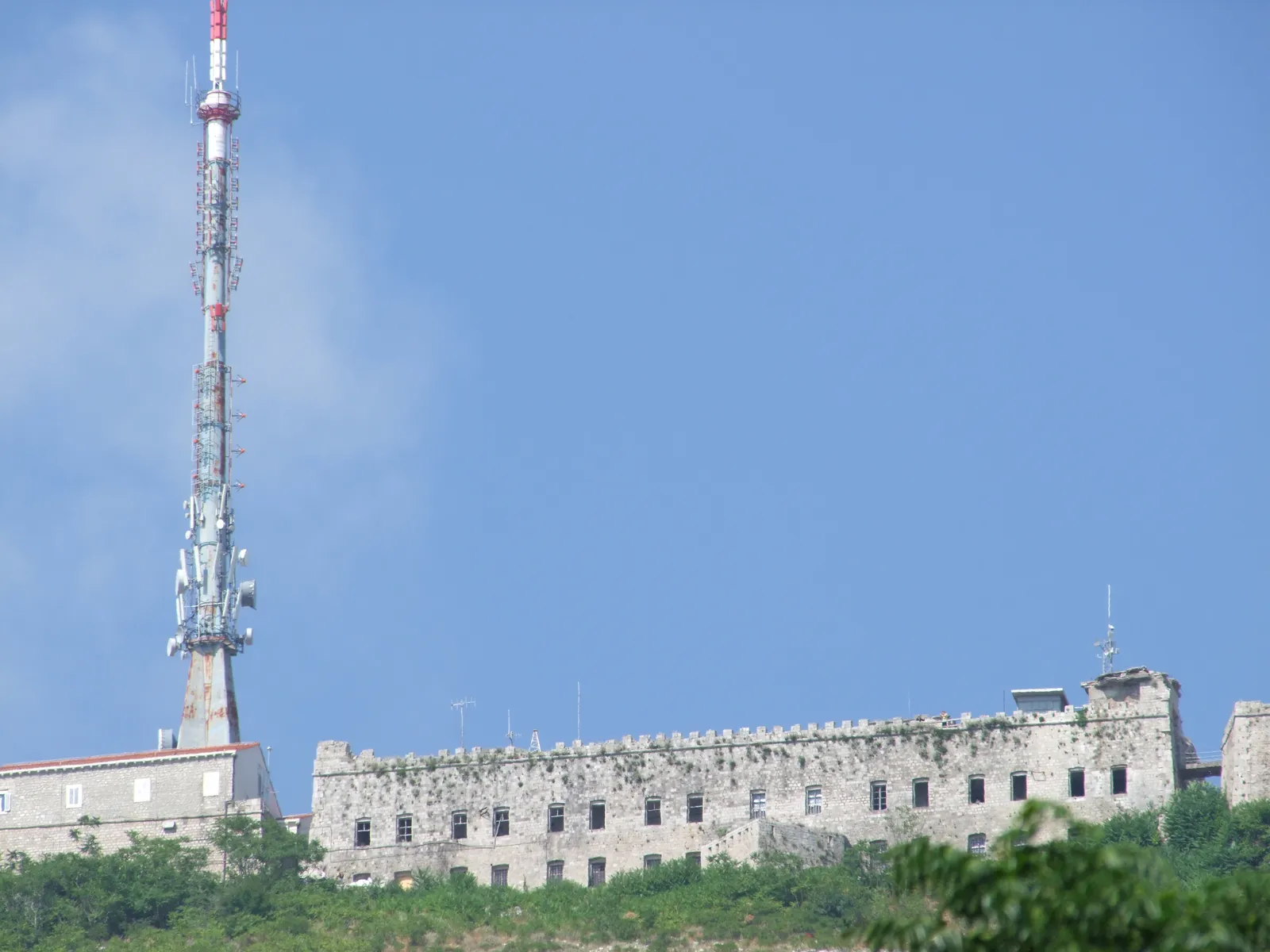 Fort Imperial along the crest of Mount Srđ, a long low crenellated stone barracks with rows of small windows, the red and white communications mast standing beside it against a clear sky