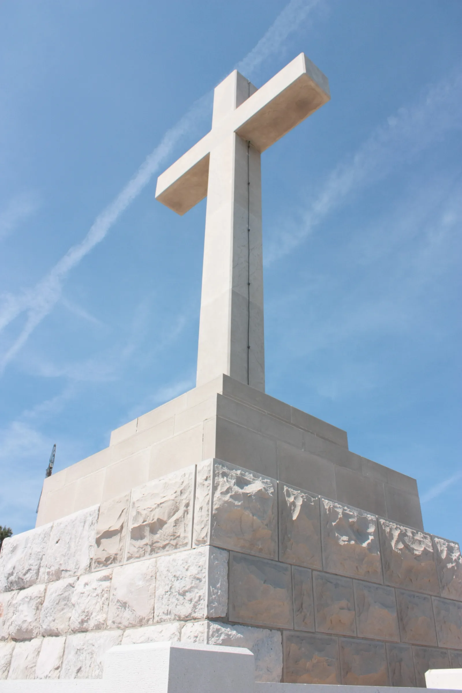 The white stone cross on Mount Srđ seen from below against a blue sky, its plain squared arms rising from a stepped block base of pale and grey limestone