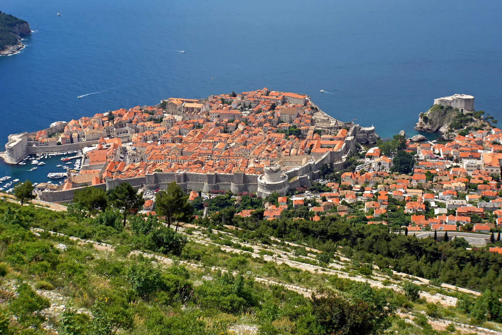 The bare scrub-covered slope of Mount Srđ dropping towards Dubrovnik, loose pale rock and low bushes in the foreground with the walled Old Town and the sea below