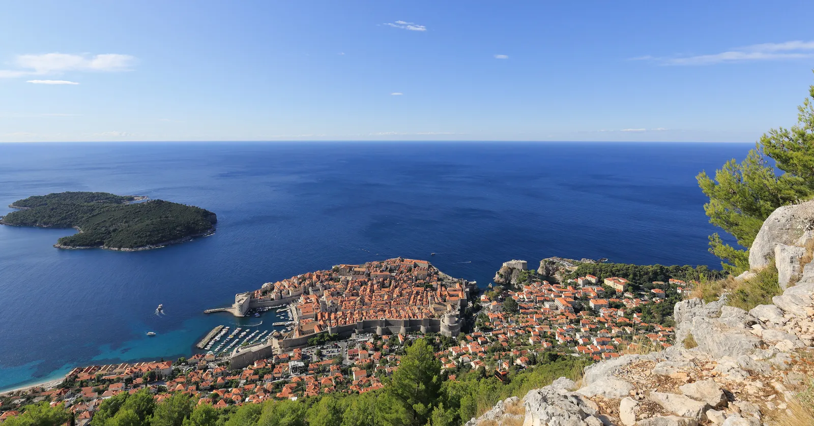 The view from Mount Srđ over Dubrovnik, the walled Old Town on its promontory with the Old Port beside it, the wooded island of Lokrum offshore and open blue Adriatic to the horizon