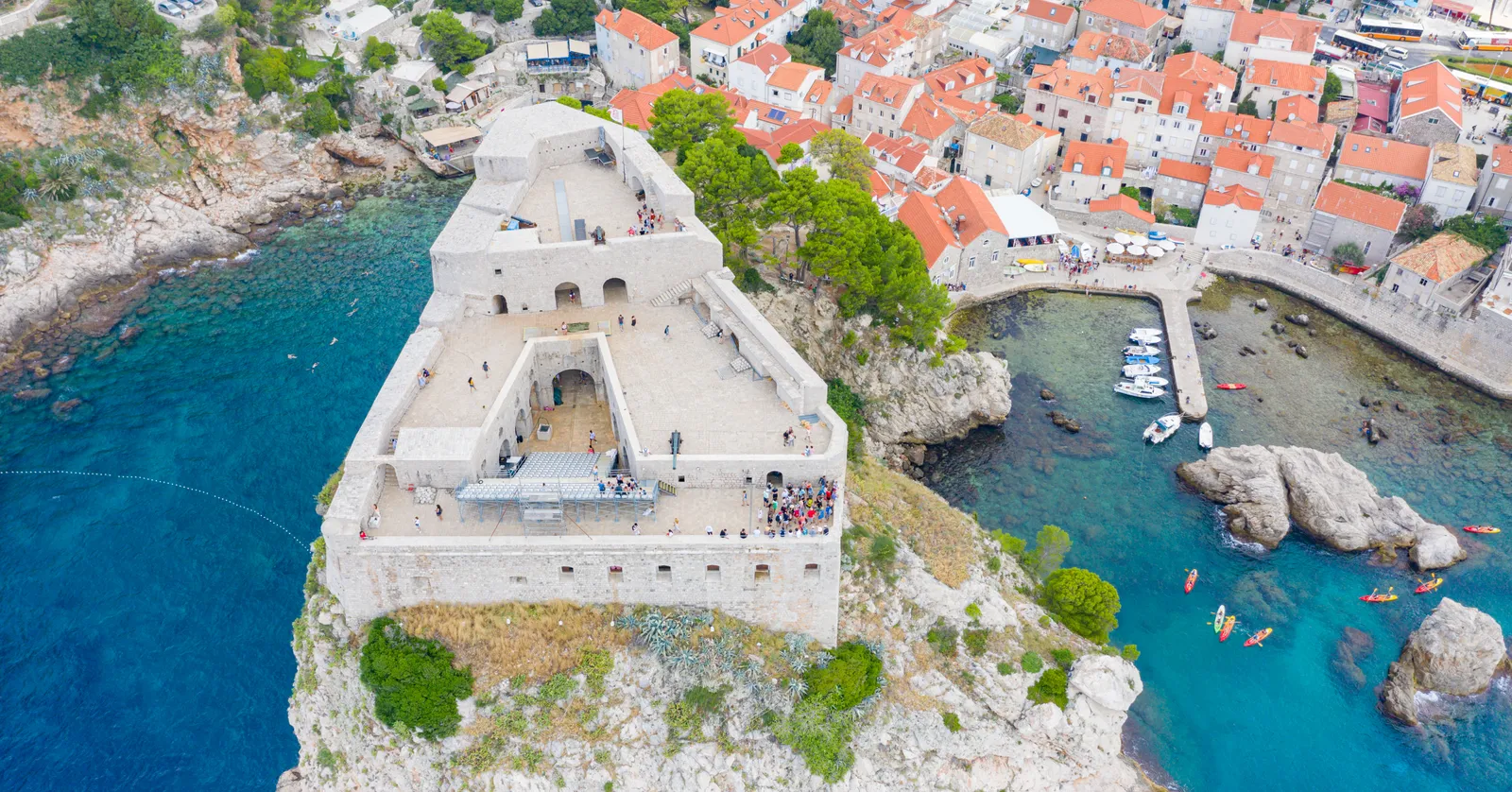 Fort Lovrijenac in Dubrovnik from the air, a square stone fortress with an open courtyard standing on a bare rock above turquoise water, with a small harbour and the orange roofs of the town behind it
