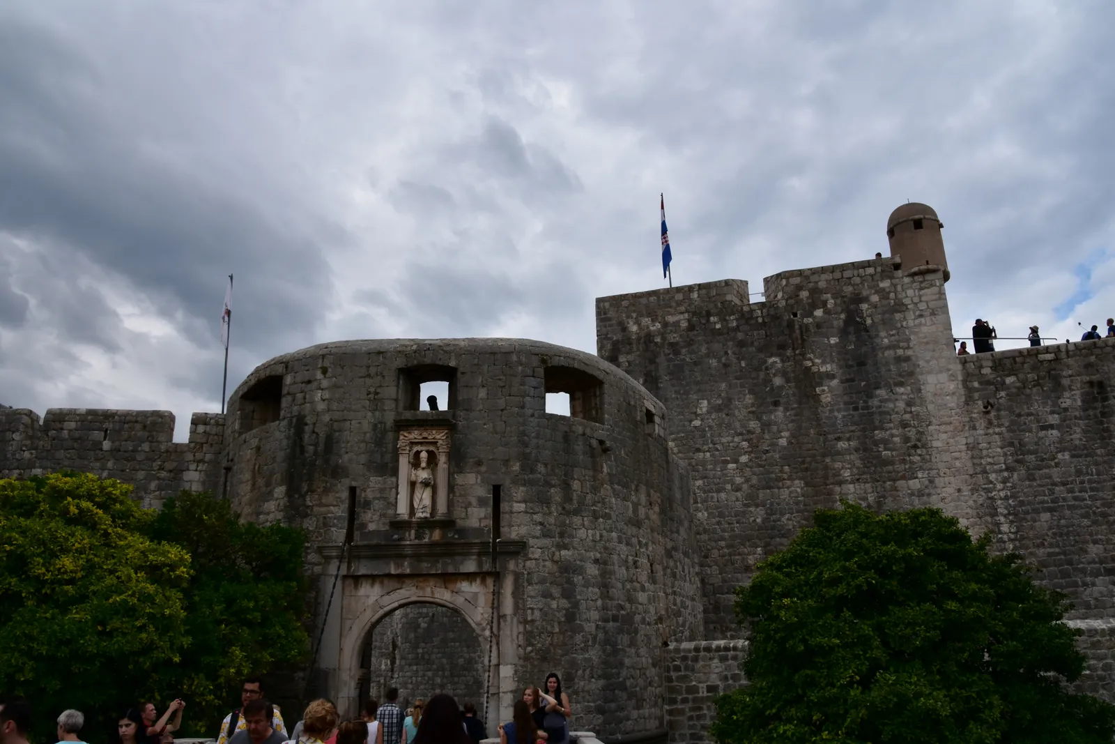 The outer Pile gate at Dubrovnik under a grey sky, a rounded stone bastion with an arched entrance and a saint's figure in a niche above it, joined to the high city wall on the right
