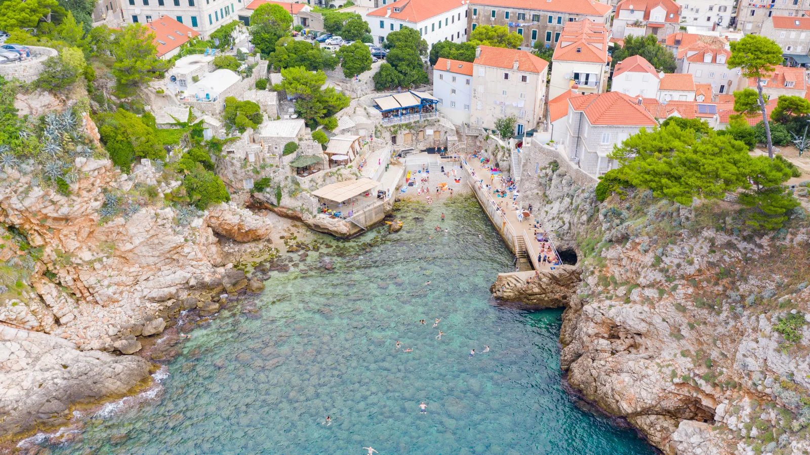 Šulić beach in Dubrovnik from above, a narrow rocky inlet of clear green water with a small pebble shore at its head, swimmers in the bay and houses stacked up the slope behind