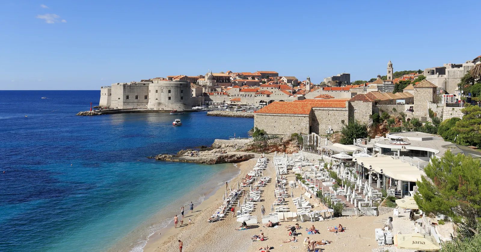 Banje beach in Dubrovnik with rows of white parasols and loungers on the pebbles, the Old Port and the round Fort St John across the water, and the walled Old Town rising behind them