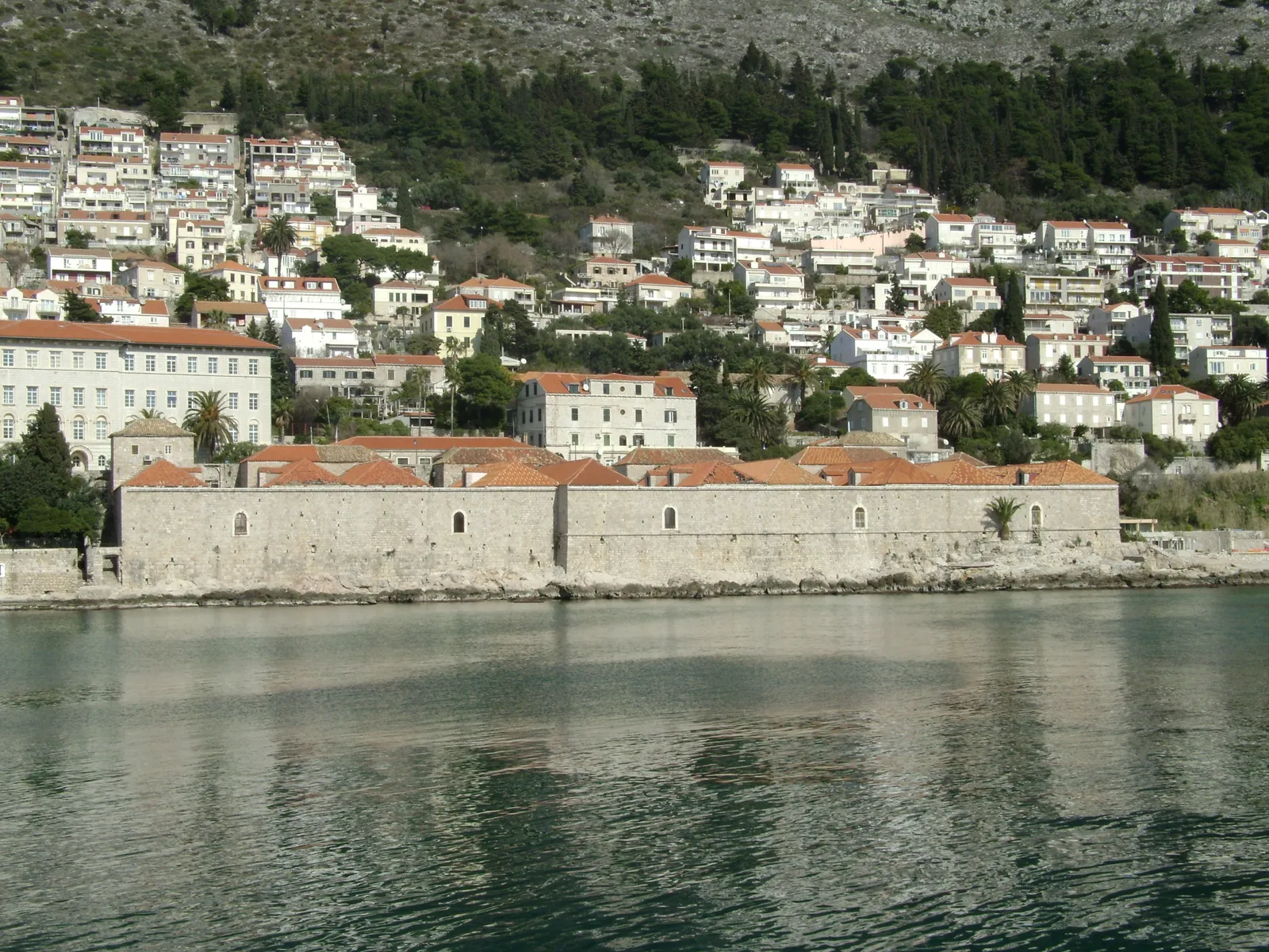 The Lazareti quarantine complex on the shore east of Dubrovnik's Old Town, a long low walled compound of terracotta-roofed blocks at the water's edge with the houses of Ploče climbing the hill behind