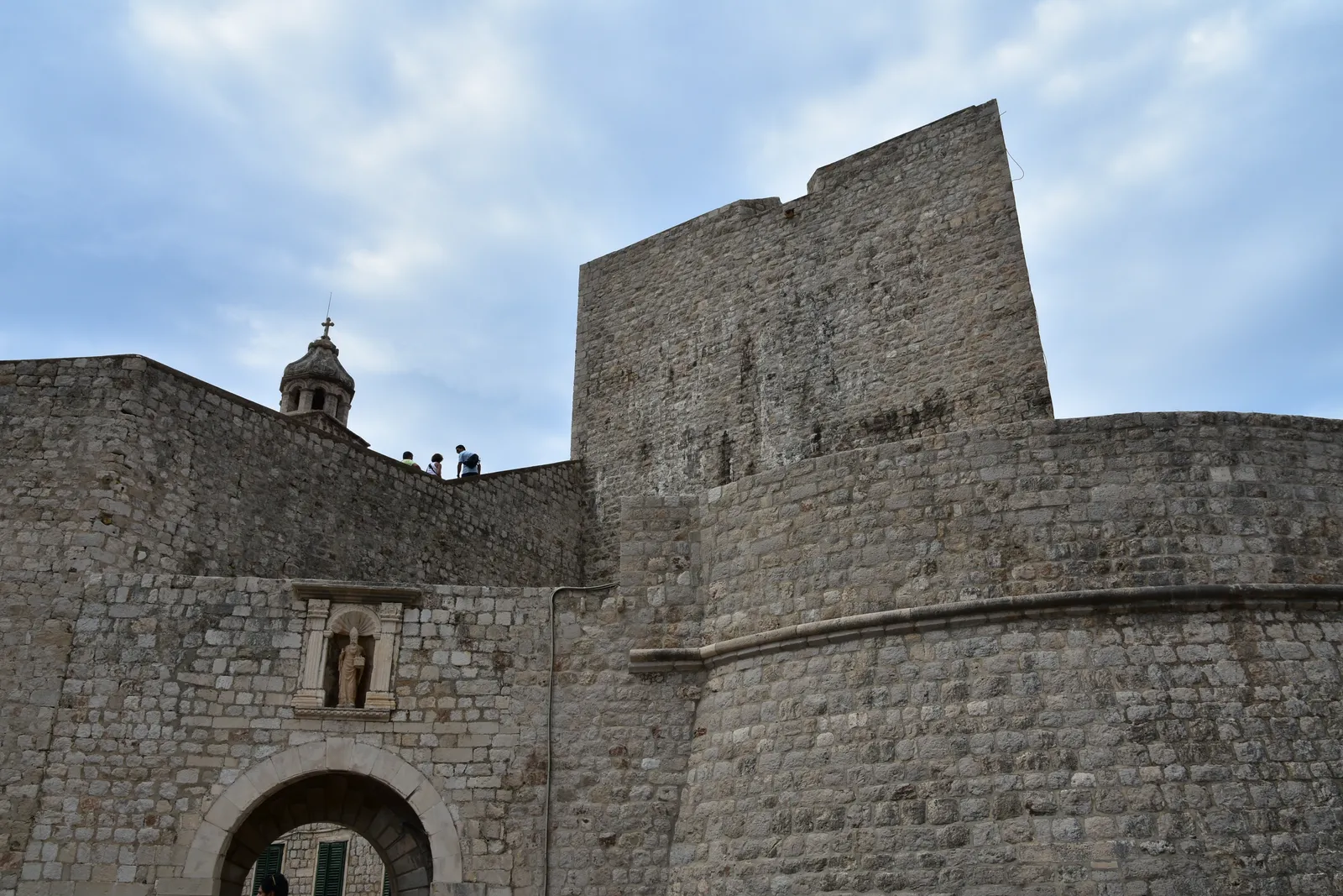 The Ploče gate on the eastern side of Dubrovnik's Old Town, a low stone arch with a saint's figure above it set into the base of a tall square bastion, with a bell turret on the wall behind
