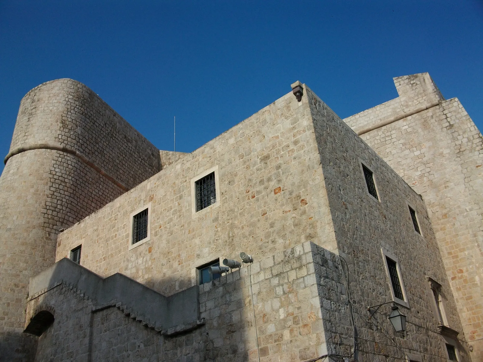 Fort Revelin in Dubrovnik seen from below against a deep blue sky, a broad block of pale stone with a rounded bastion on one corner and small square windows high in the wall
