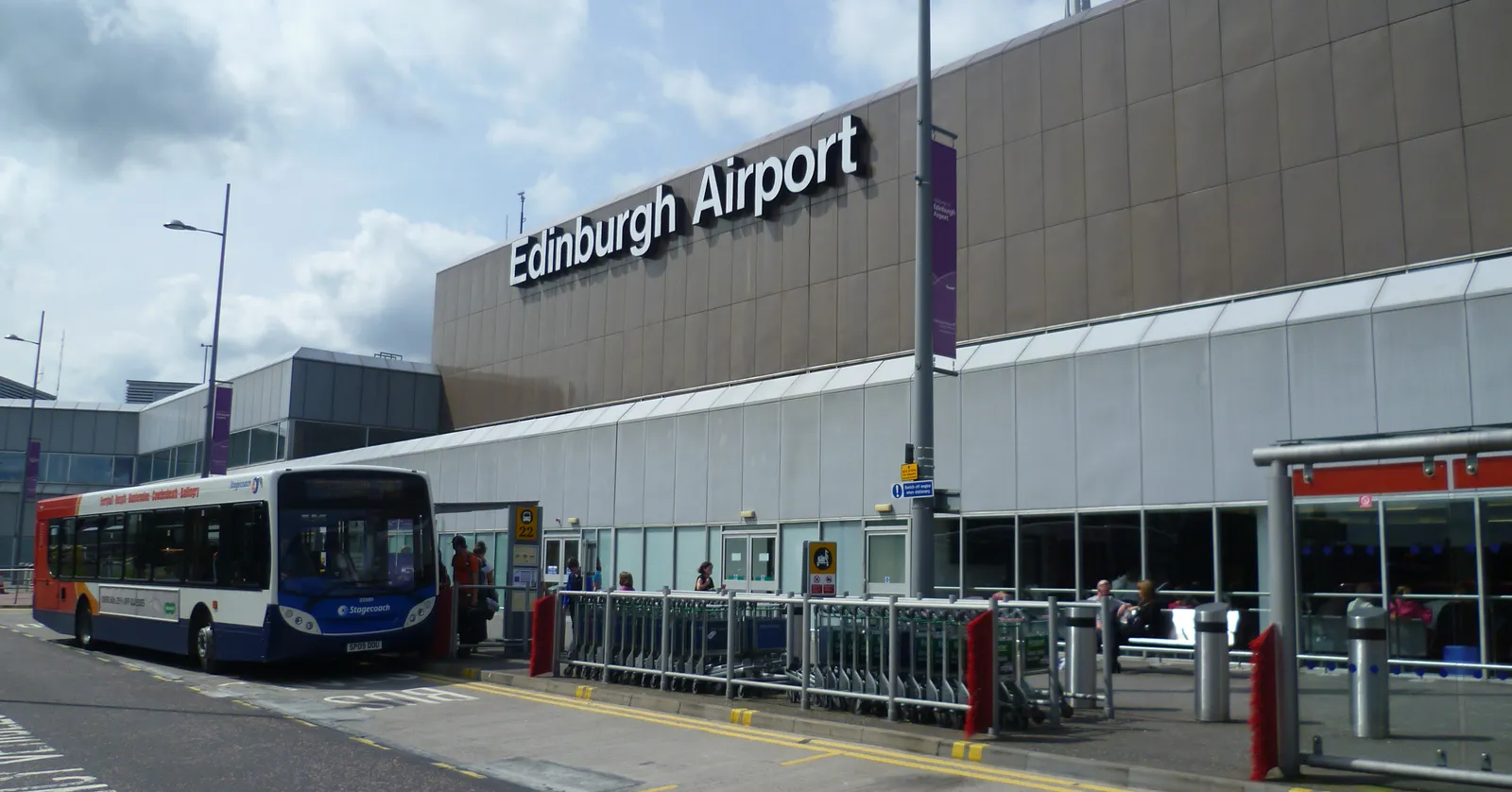 The frontage of Edinburgh Airport's single terminal, its name in large letters above the departures doors, with a bus pulled up at the kerb outside