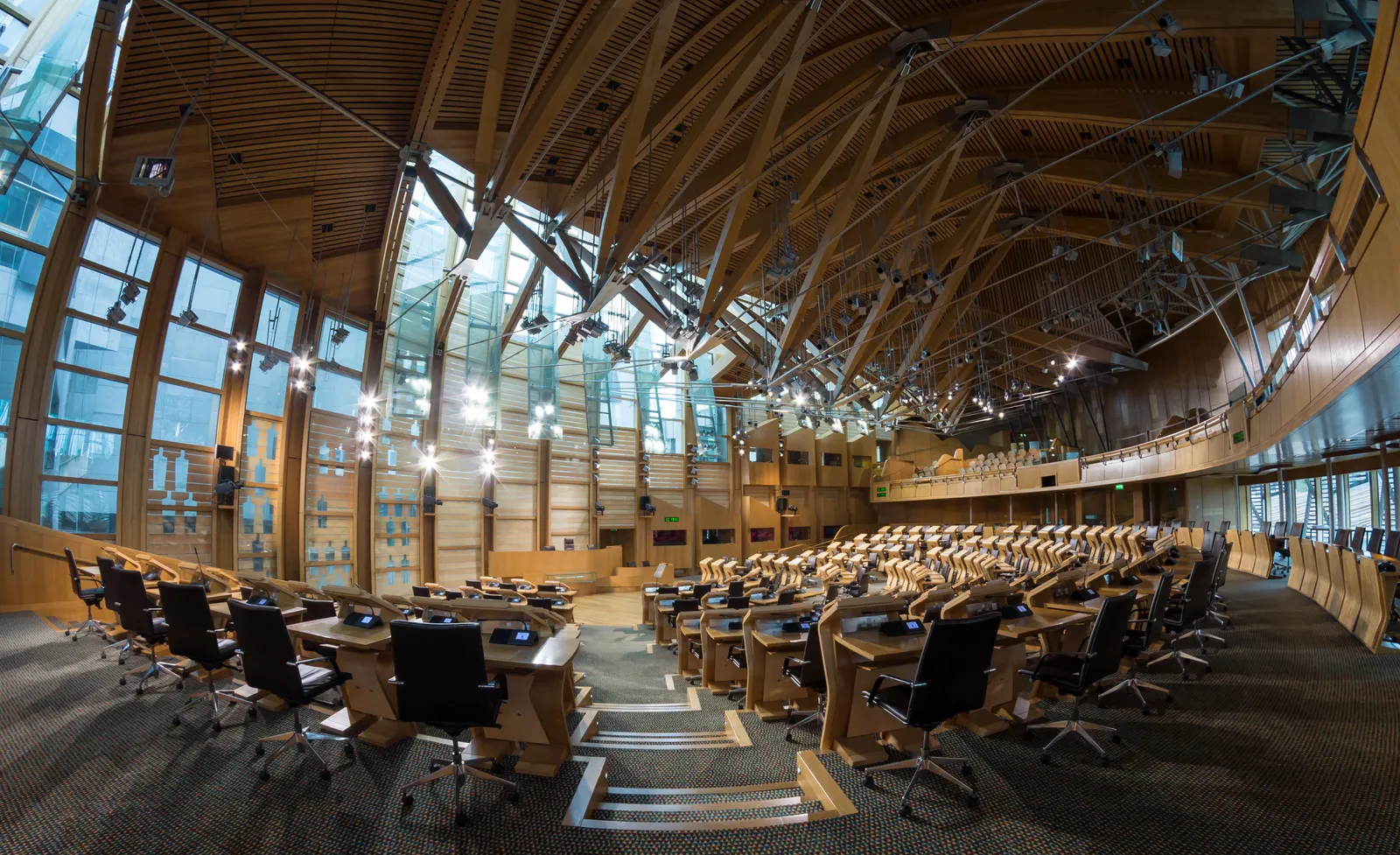 The debating chamber of the Scottish Parliament, curved rows of desks under a roof of laminated oak beams and stainless steel struts with glass between them