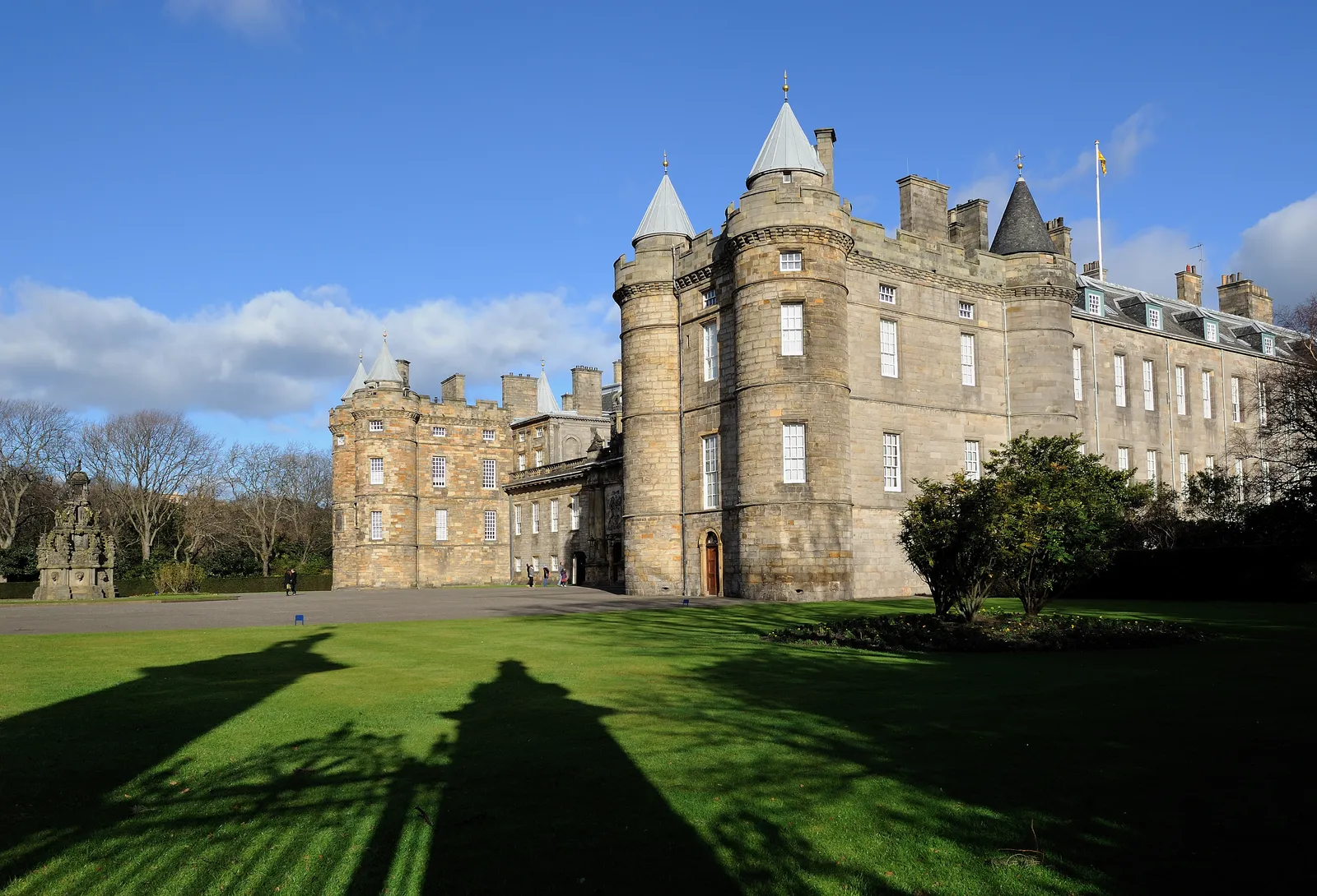 The Palace of Holyroodhouse in Edinburgh, its turreted west front and forecourt in low sun, with the ruined gable of Holyrood Abbey visible at the left