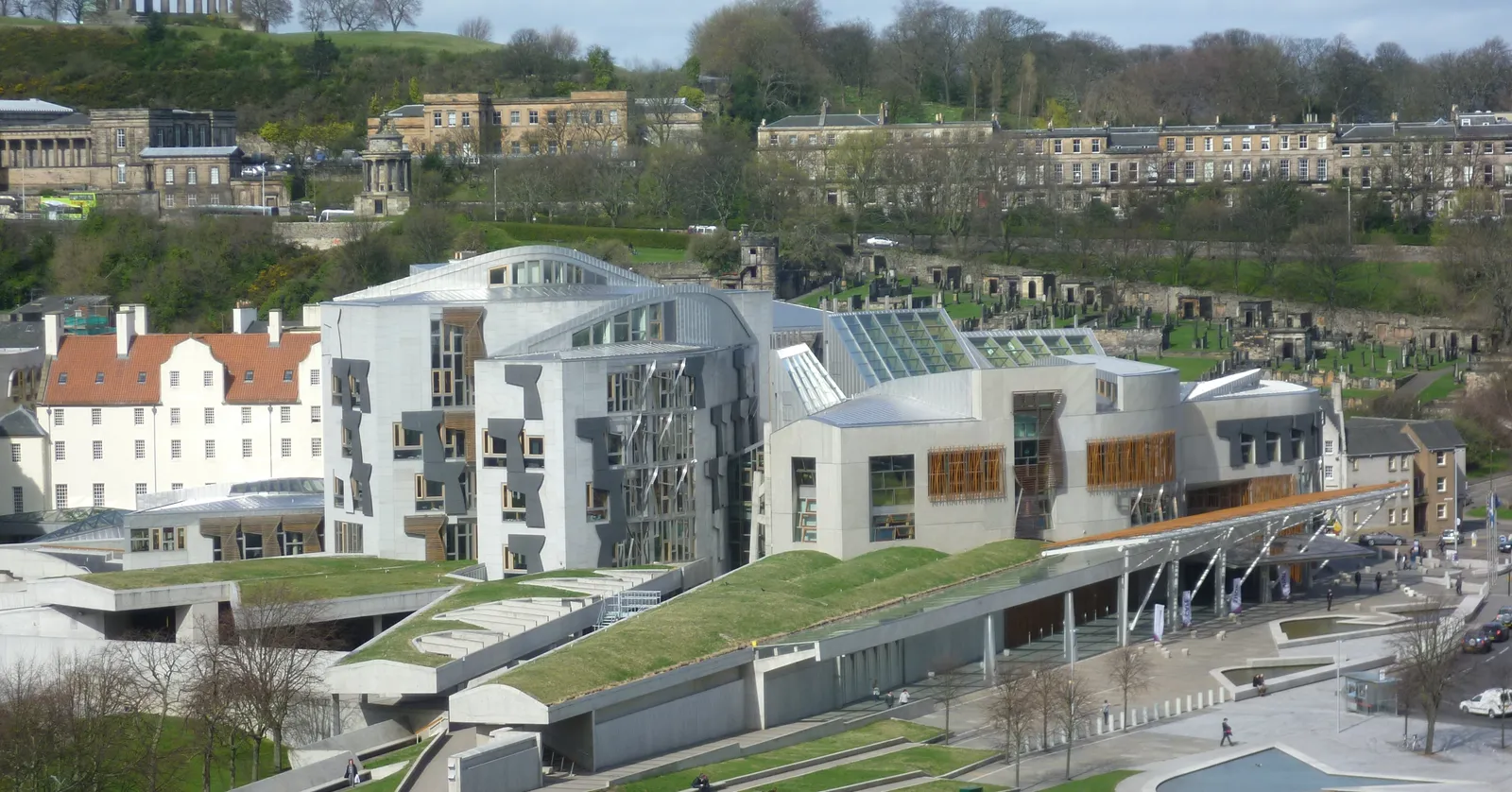 The Scottish Parliament complex at Holyrood in Edinburgh seen from above, its low grass-roofed wings and angular concrete and oak facades set against the Georgian terraces behind