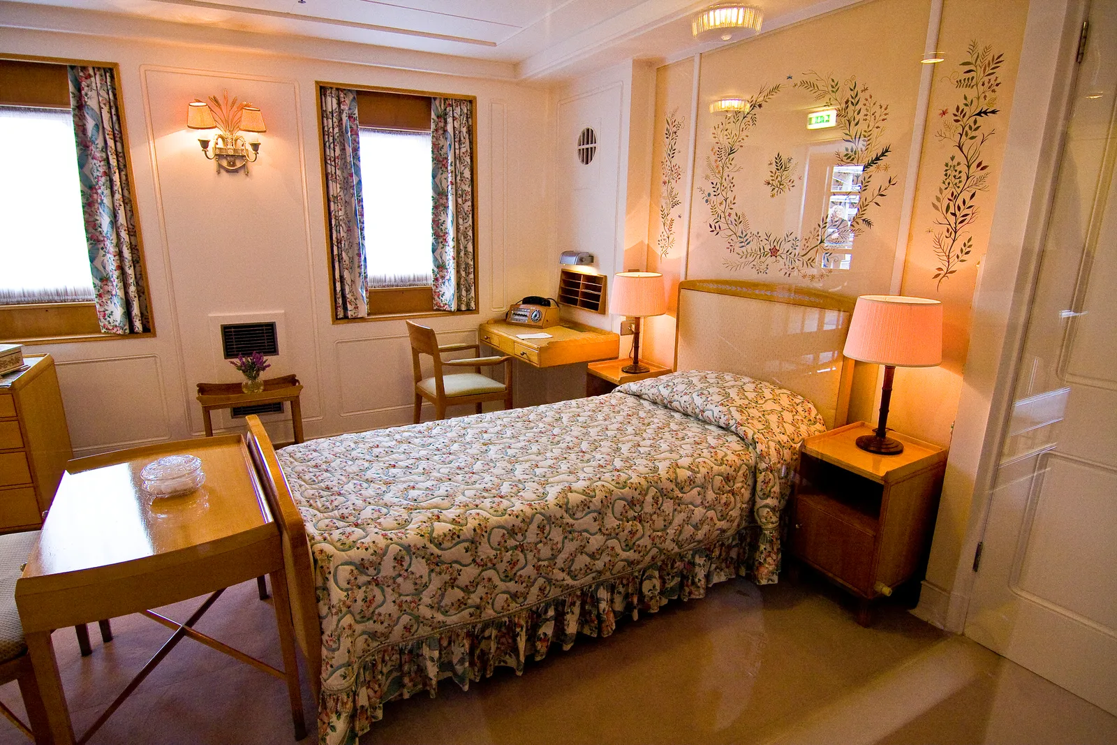 A cabin aboard the former royal yacht Britannia, with a single bed, a floral quilt, wall lamps, a writing desk and two portholes looking out over the water