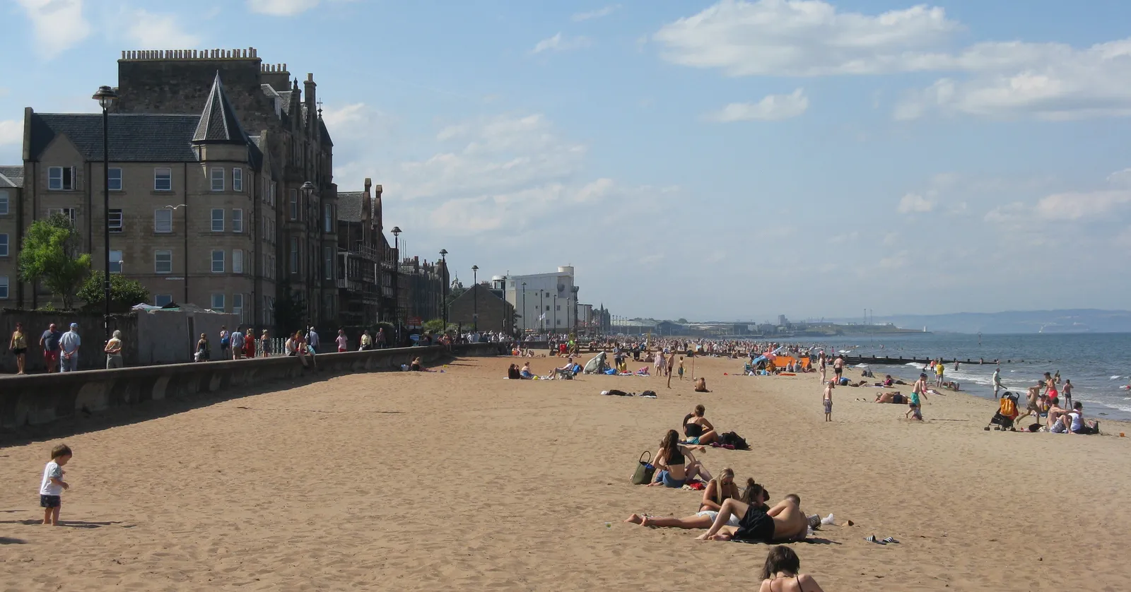 The sand at Portobello in Edinburgh on a sunny day, people sitting along the beach with the tenements and sea wall of the promenade running behind them