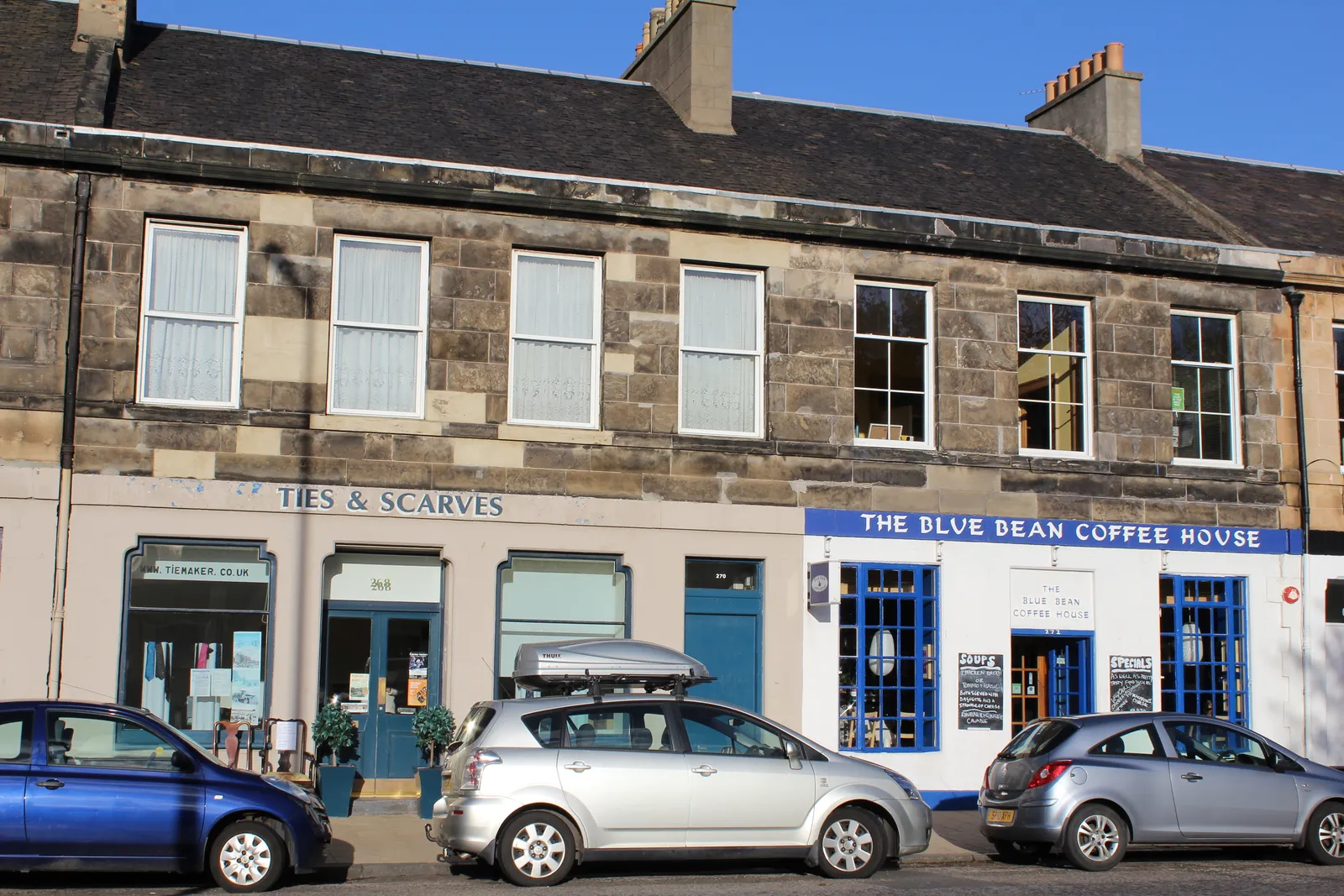 A terrace of stone buildings on Portobello High Street in Edinburgh, with a coffee house and small independent shopfronts at street level and cars parked outside