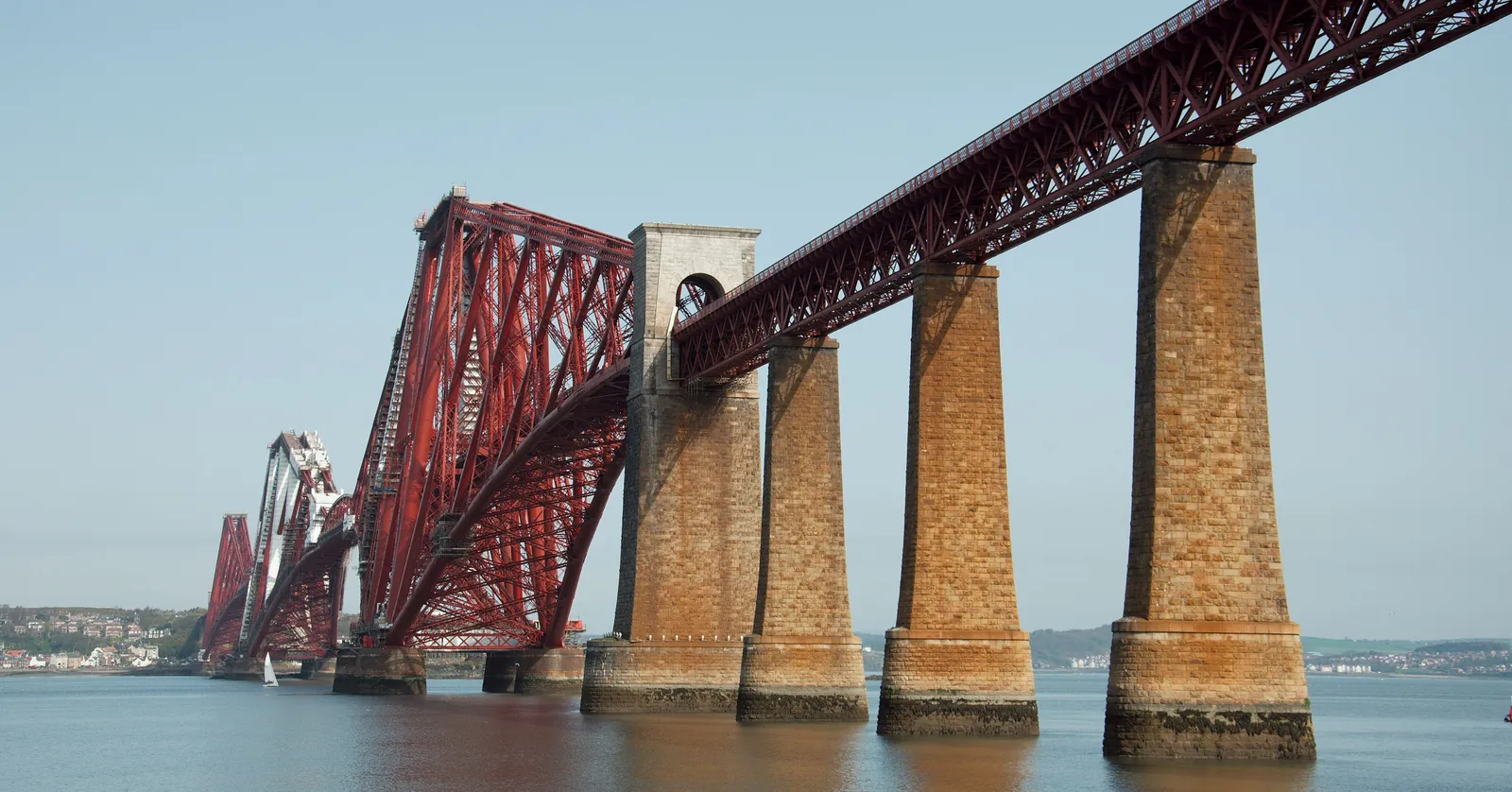The Forth Bridge seen from below at South Queensferry, its red steel cantilevers carried on tall stone piers standing in the water of the firth