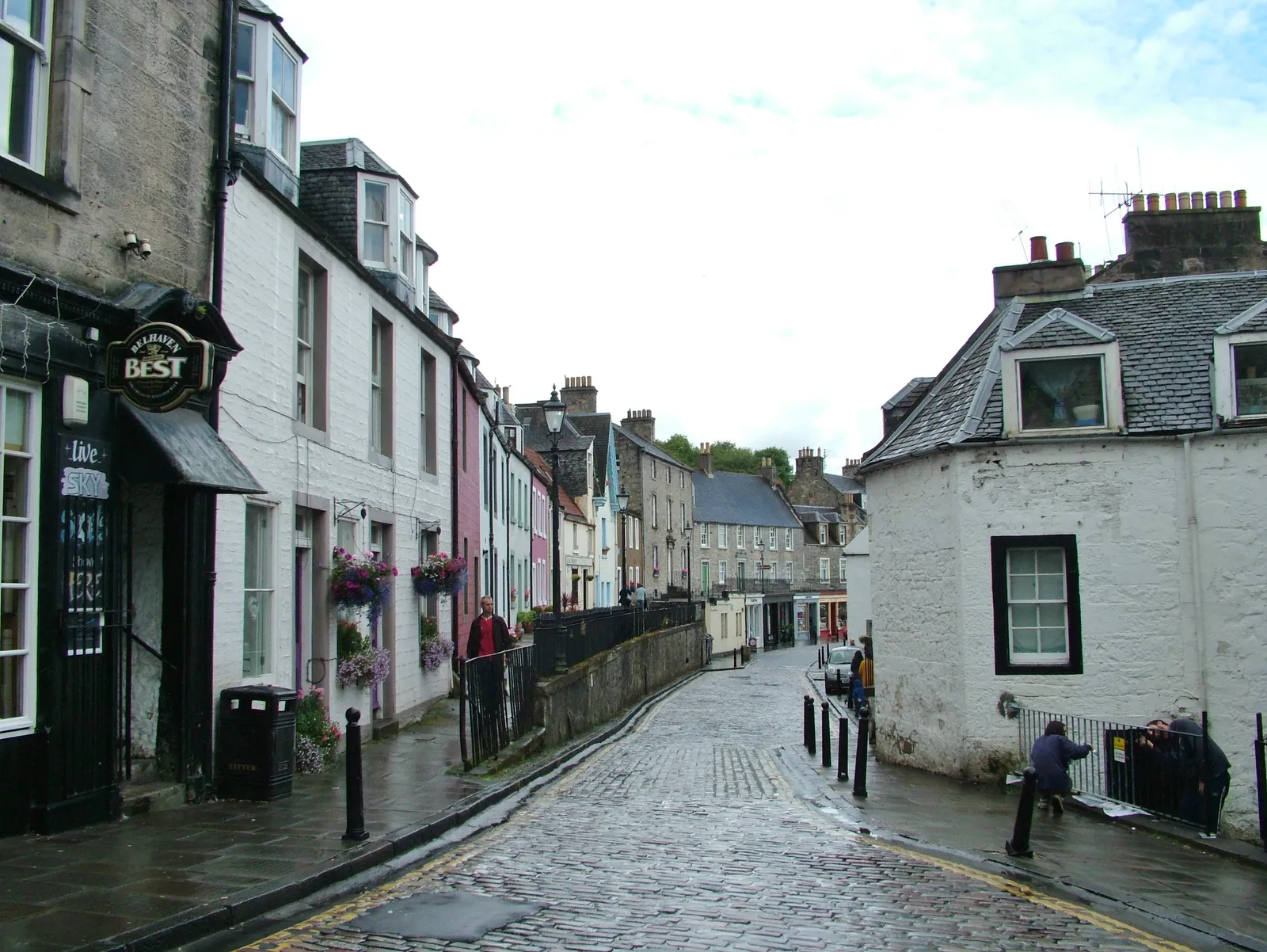 The cobbled High Street in South Queensferry, narrow and wet, lined with painted stone houses and a raised terrace of buildings on the right