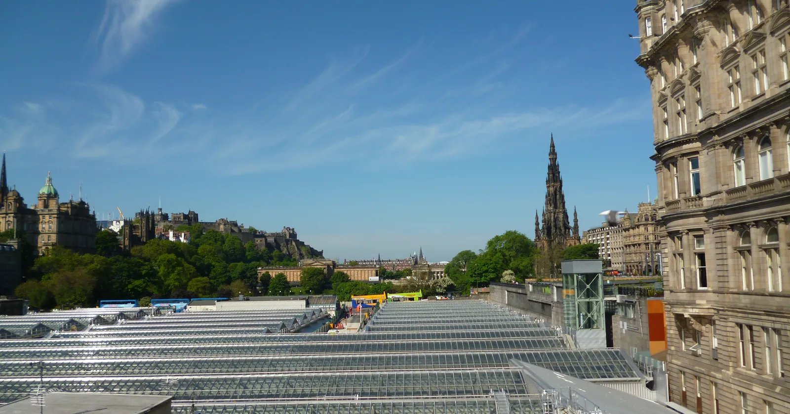The long glass ridge-and-furrow roof of Edinburgh Waverley station filling the valley below Princes Street, with the Scott Monument's dark spire on the skyline behind it