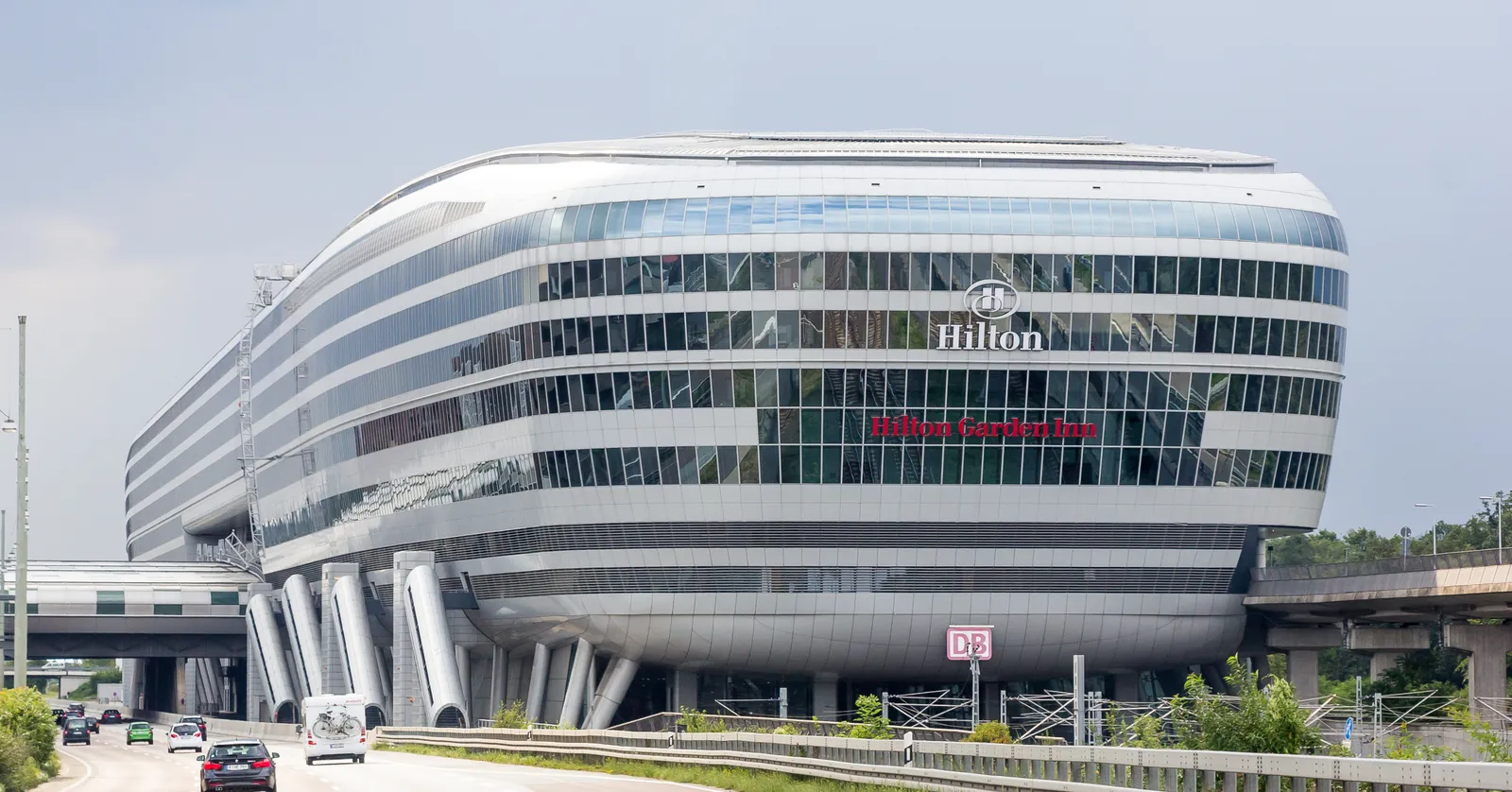 The Squaire, a long curved glass building on stilts straddling the motorway and rail tracks at Frankfurt Airport, with a Hilton sign on its facade