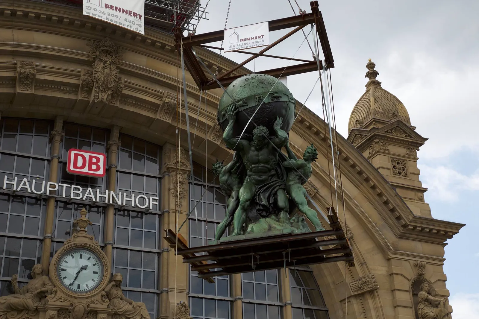 The bronze statue of Atlas holding a globe above his shoulders, suspended on a crane platform against the Hauptbahnhof clock and facade during its 2014 restoration