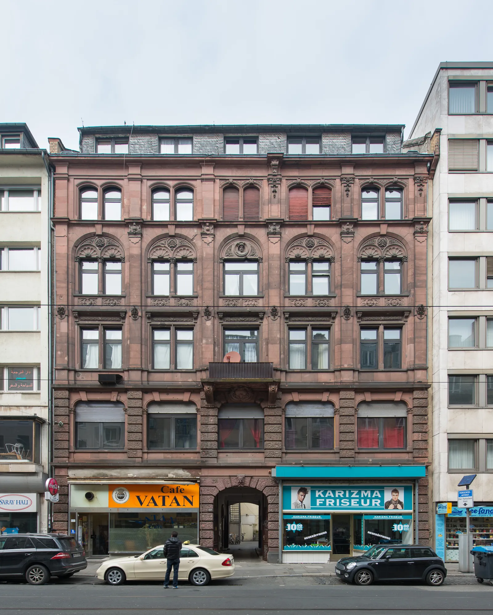 A four-storey red sandstone Gründerzeit building on Münchener Straße with a Turkish café and a barber shop at street level and parked cars along the curb