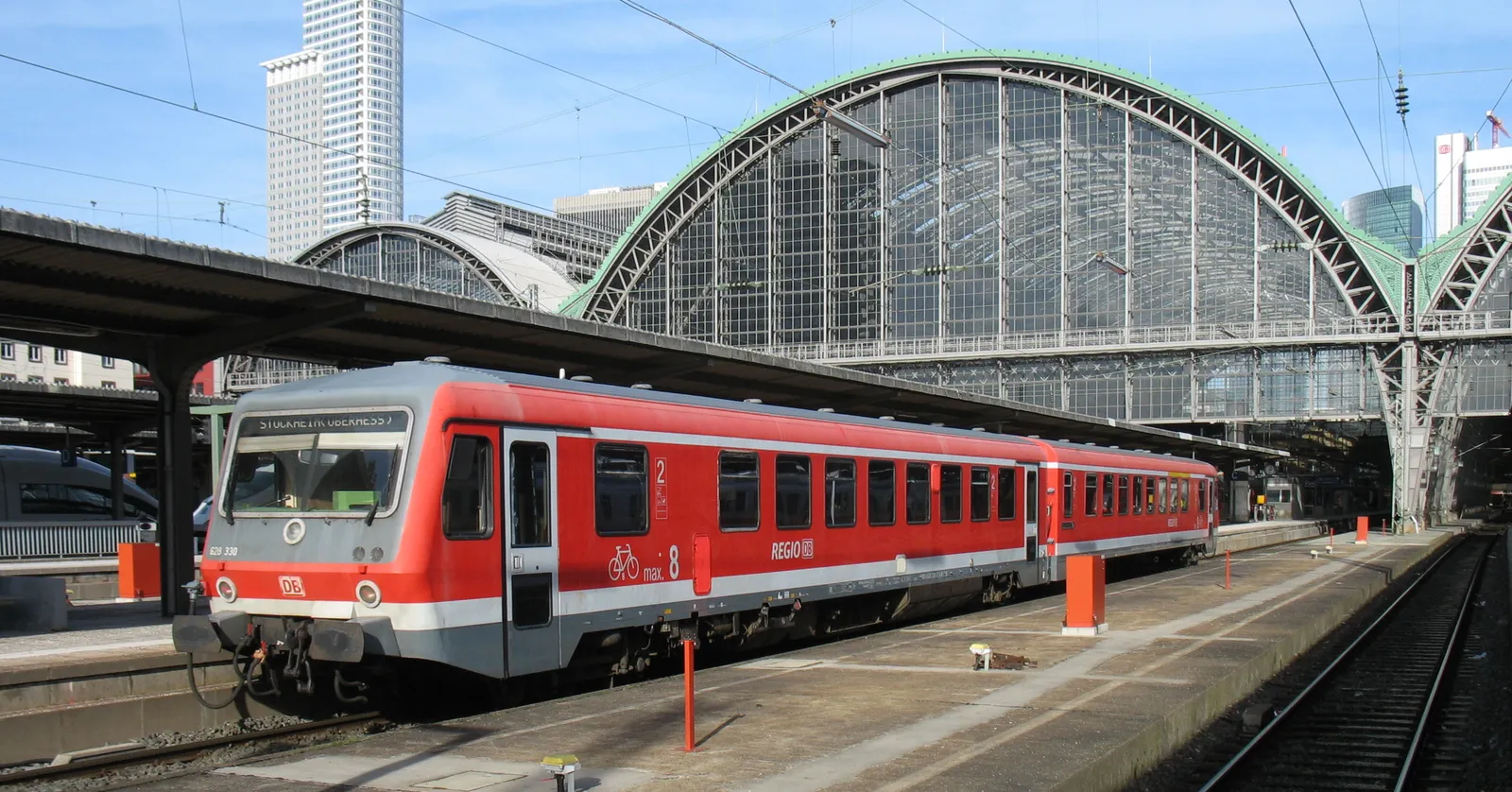 A red regional train waiting under Frankfurt Hauptbahnhof's arched iron-and-glass train shed, with the city's bank towers rising behind it