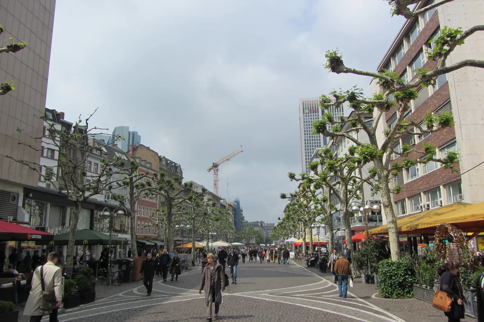 A pedestrianized street in Frankfurt's Fressgass lined with trees, café awnings and market stalls, with shoppers walking down the middle