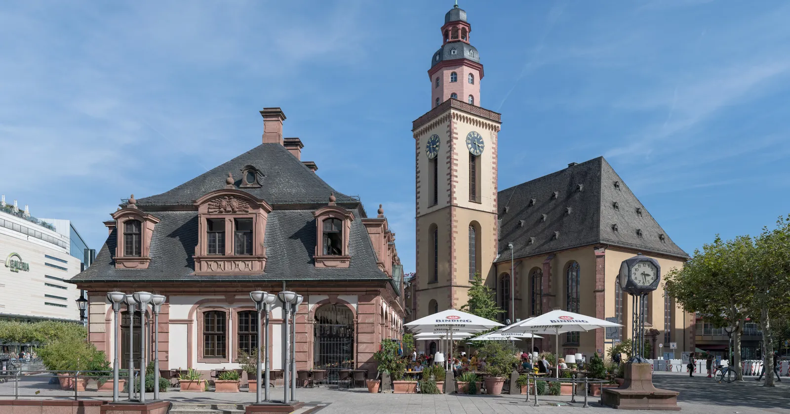 The pink-and-cream baroque Hauptwache building with its steep roof and café tables out front, beside the taller tower of Katharinenkirche church on a sunny square in Frankfurt