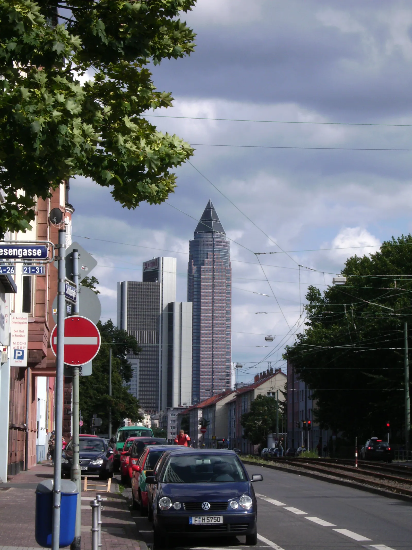 The Messeturm's tapered, pencil-shaped glass tower rising above Frankfurt's Westend district, seen down a tree-lined street