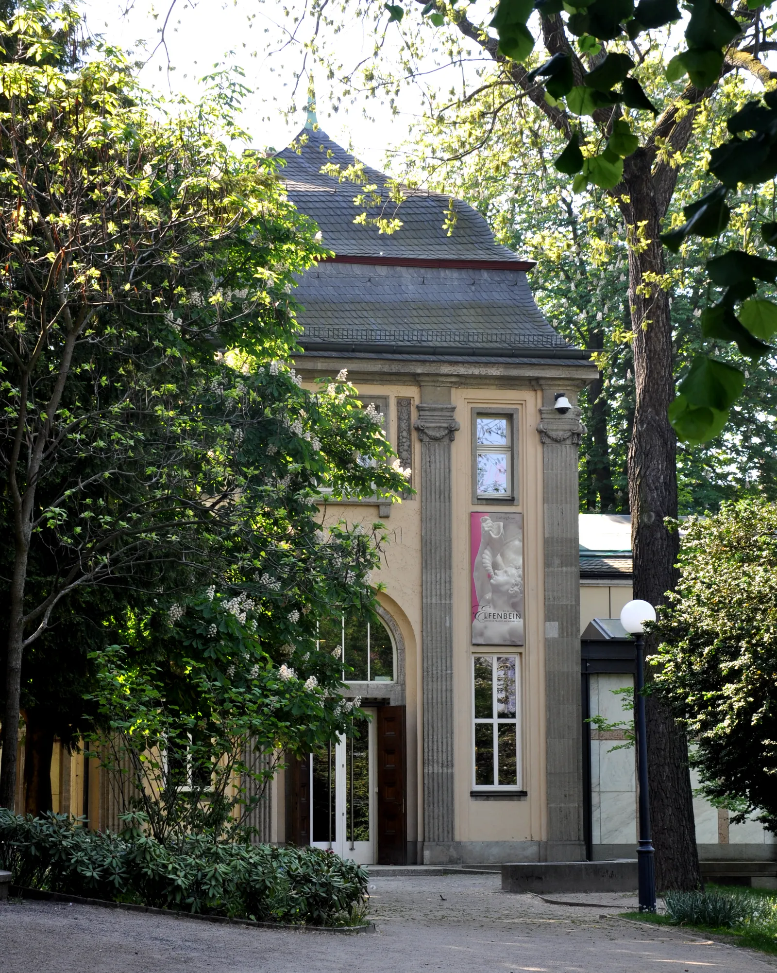 The Liebieghaus's turreted 19th-century villa entrance in Frankfurt, framed by garden trees with a sculpture visible through a tall arched window